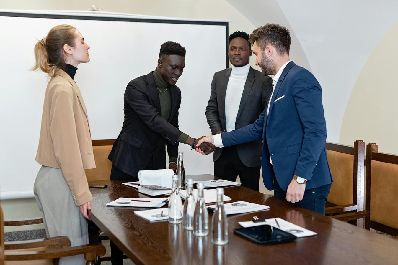Four professionals in a modern office, two men shaking hands signifying agreement.